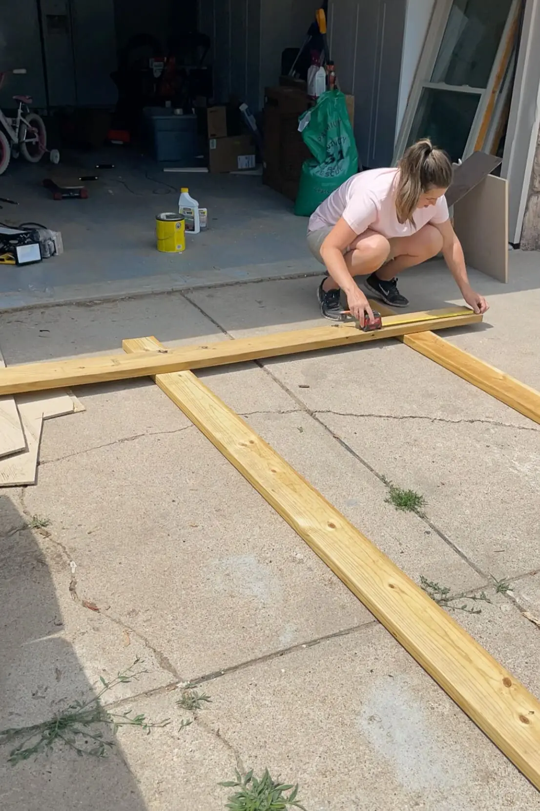 A woman wearing a pink shirt and black sneakers crouches on a concrete driveway, measuring and marking a wooden beam with a red tape measure. The project is laid out with multiple wooden planks, forming the base structure of a woodworking project. In the background, an open garage with tools, supplies, and a child’s bicycle is visible.